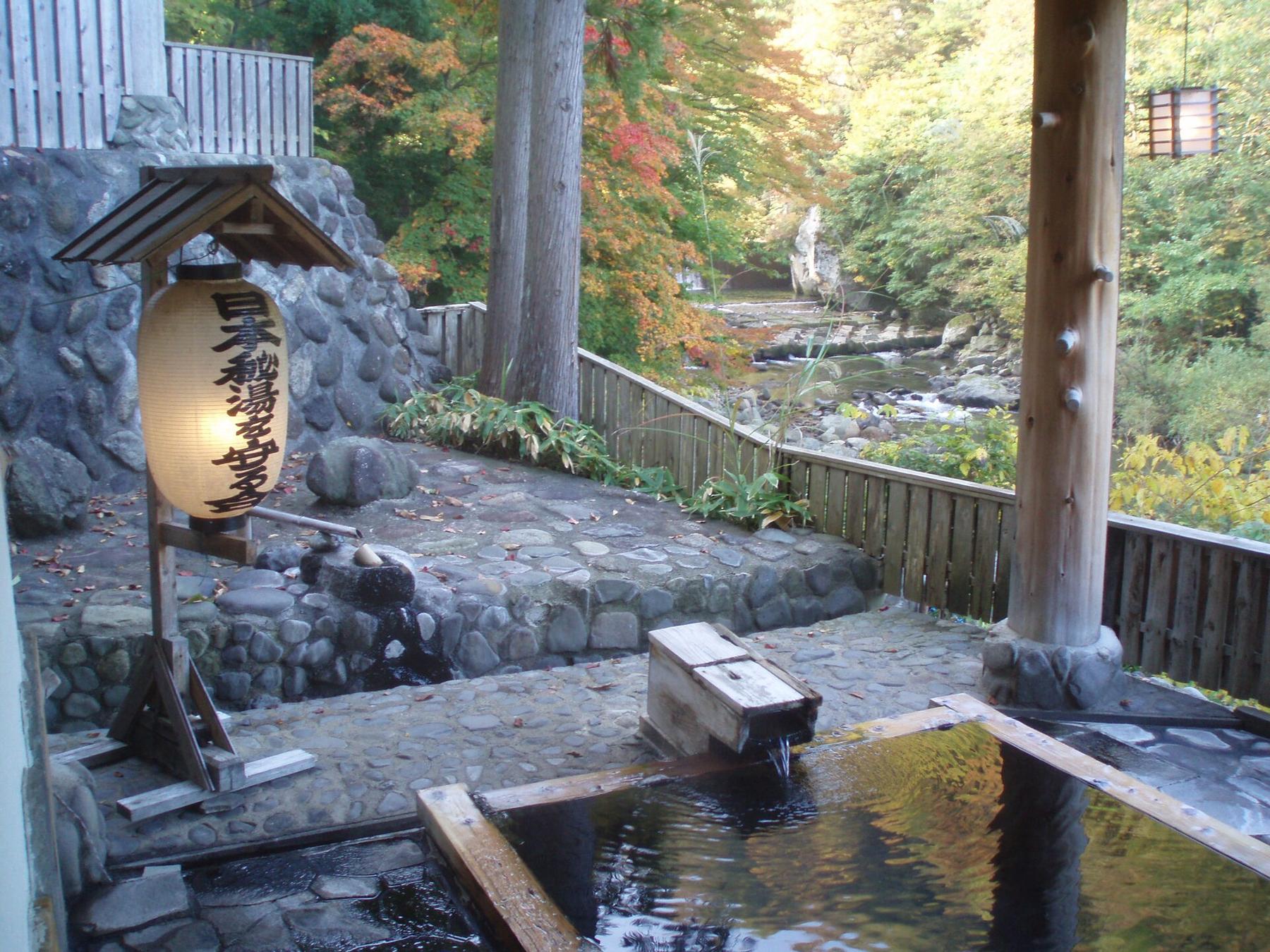 An open-air hot spring bath beside a stream, with a lantern and timber posts